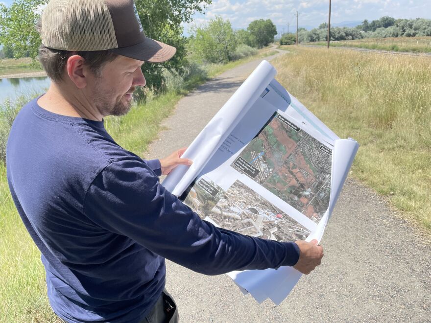 Boulder County Parks and Open Space planner Justin Atherton-Wood holds open a scrolled piece of white paper with images on it while standing on a dirt path with green grasses on either side.