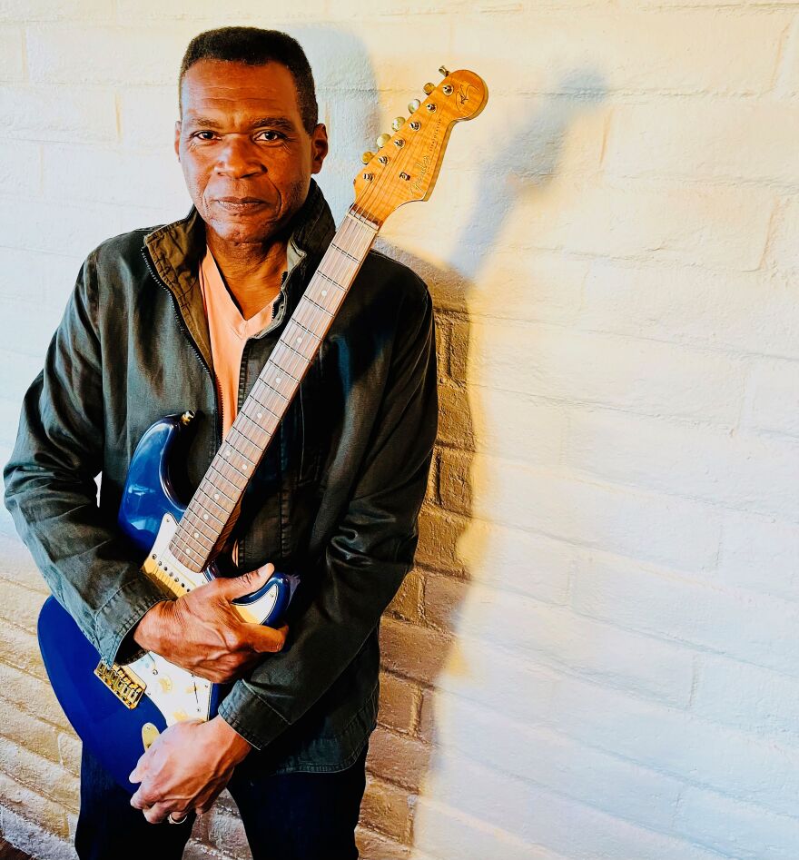 Leaning against a wall, Robert Cray holds a blue electric guitar.