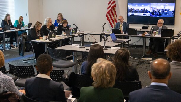 A group of people in business attire sit at tables arranged in a U-shape, attending a meeting with lawmakers in a conference room. An American flag is visible, and a video conference is displayed on a screen in the background.