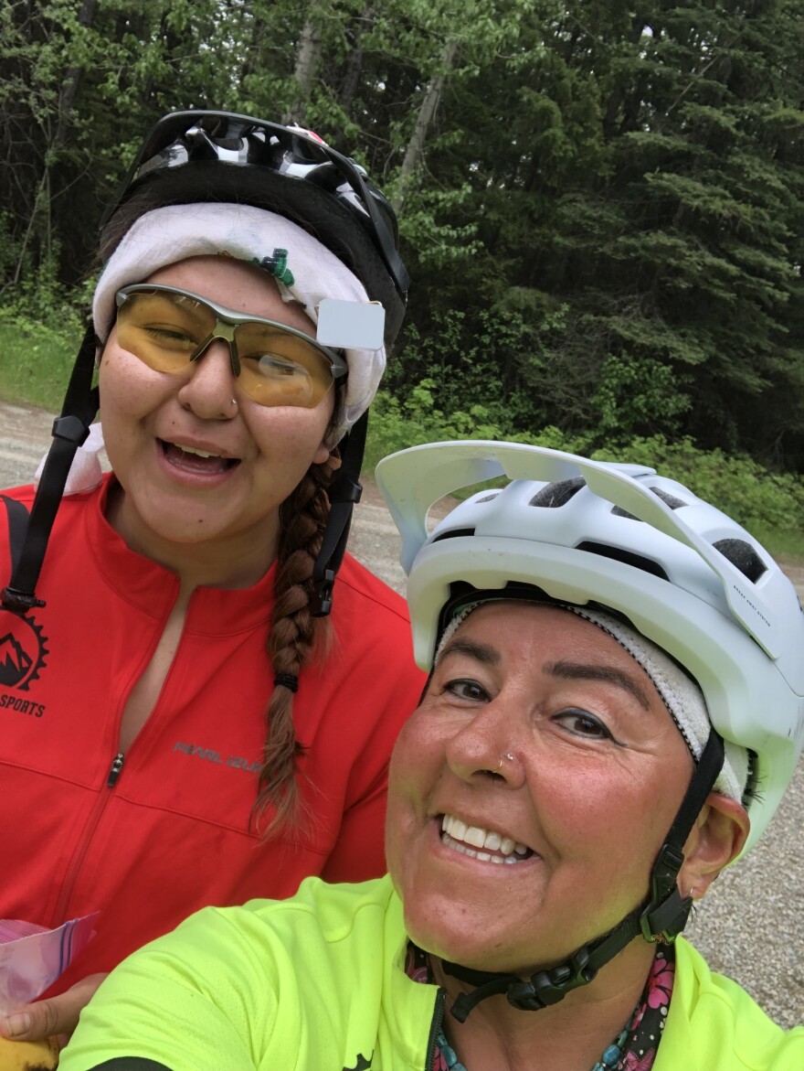An older woman in a white bike helmet and bright yellow shirt takes a selfie with a younger woman in a red shirt and a bike helmet.