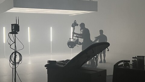 Two silhouetted crew members operate a mobile camera rig in a hazy studio. 