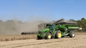 A green John Deere tractor pulling a wagon drives next to a combine harvesting soybeans.
