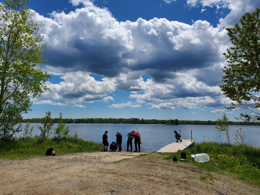 Six people on the bank of a forest lined lake near a dock under a partly cloudy sky