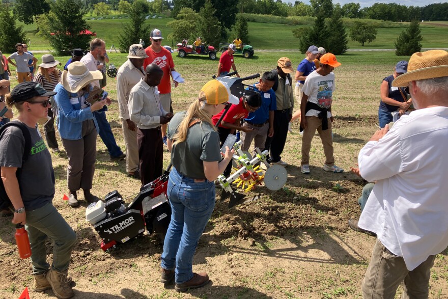A group of farmers in hats in boots stand in a dry field around a small, two wheel tractor.