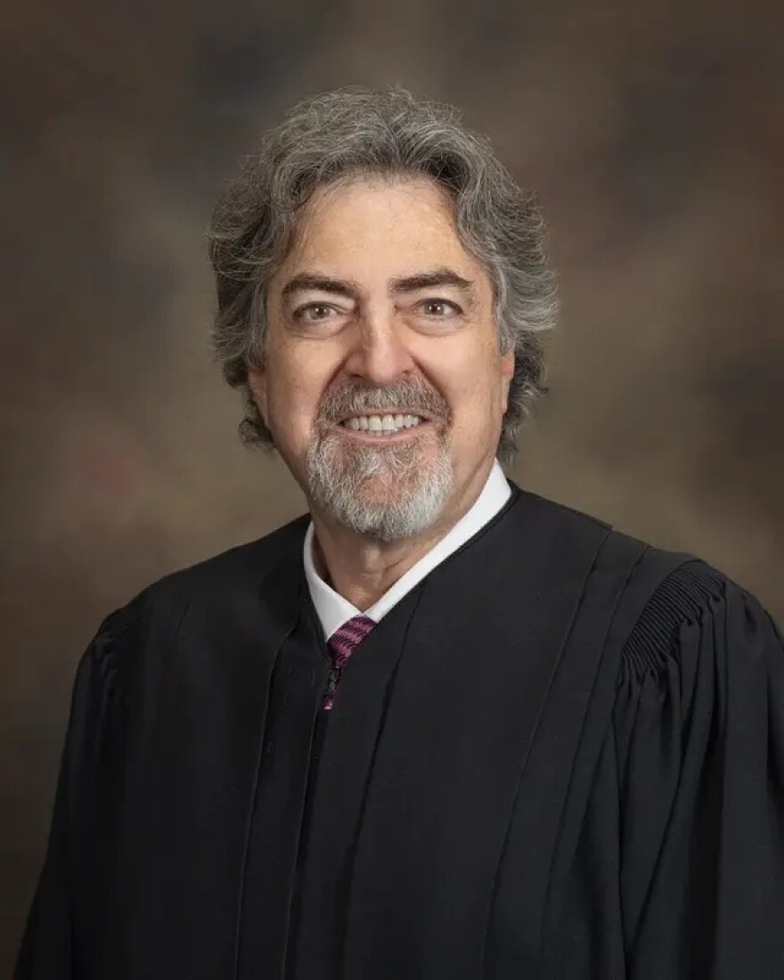 man with grey full beard, mustache and hair sits in a judge's robe
