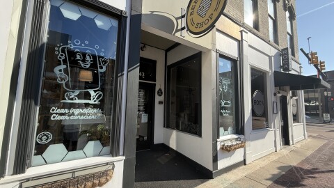The front of Bee Joyful shop is depicted on the Kalamazoo Mall. The sidewalk in front of the store is red brick, and the storefront has two large glass windows with murals. One mural is of a cartoon bar of soap with a face and hands that is waving. Underneath it are the words “clean conscience”. The Bee joyful shop sign is a yellow circle with a bee hive in the middle.