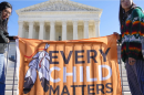 Two indigenous people stand outside the Supreme Court with a flag that has two feathers tied together and the words "Every Child Matters" on an orange background.