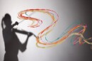 The silhouette of a violinist is cast upon the left side of a screen, created by a single spotlight. Colorful painted streaks of yellow, pink, orange, red blue are twisting and weaving across the image, originating from the violin's shadow and traveling to the right.