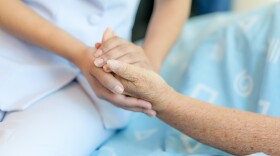 A young woman sits at the bedside of an elderly woman. The young woman holds the elderly woman's hand.