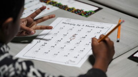 A childs hand holds a pencil as they review a school worksheet. Their other hand holds up three fingers.
