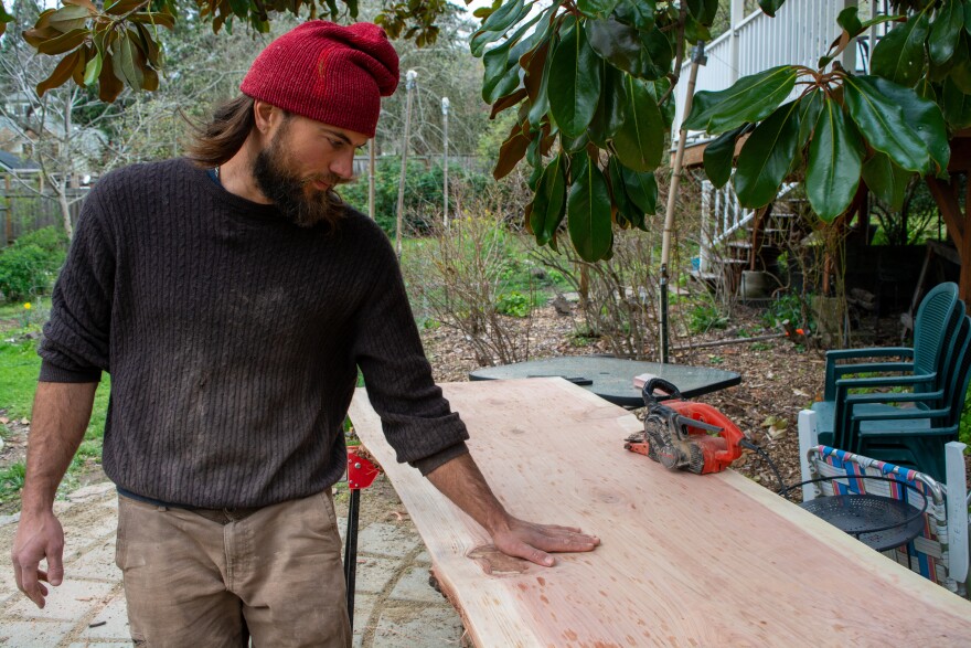 A man with a beard and medium-length hair is wearing a black sweatshirt and red beanie. His left hand is rubbing along a large slab of redwood. there's an electric sander sitting on the slab