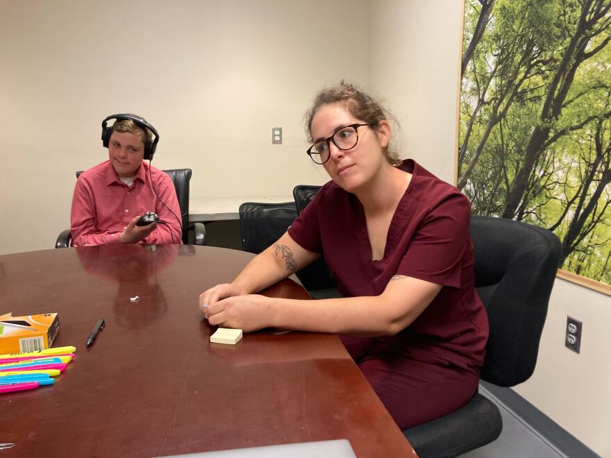 Jack sits at a dark wood table with a microphone and headphones next to a woman wearing red scrubs. There are highlighters and sticky notes on the table.