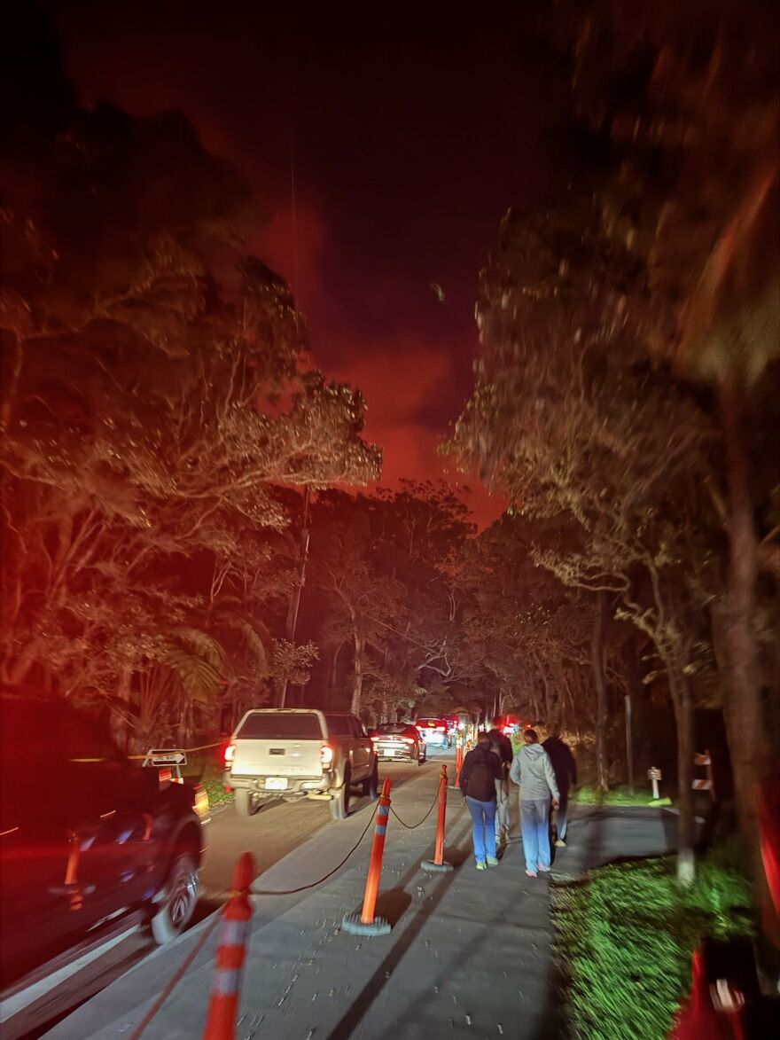 Cars drive through a single-lane detour in Hawaiʻi Volcanoes National Park, with visitors walking alongside the vehicles separated by cones and rope. The sky is red from the lava erupting out of Kīlauea volcano.