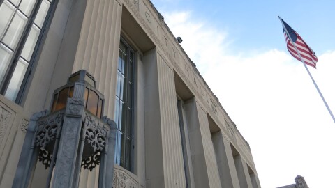 Kalamazoo city hall is seen from below. It is a large grey stone building with intricate carved details, including a hand-carved "city hall" on the front of the building