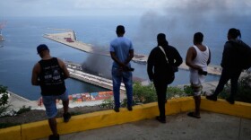 Men watch smoke rising from a dock after explosions were heard at La Guaira port, Venezuela, Saturday, Jan. 3, 2026.