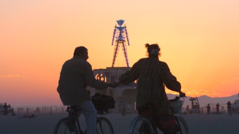 A couple holds hands while cycling near The Man, a stick figured symbol of the Burning Man art festival, during a morning sunrise Saturday, Sept. 3, 2006, at the 20-year-old art festival in Nevada's Black Rock Desert. (AP Photo/Ron Lewis)