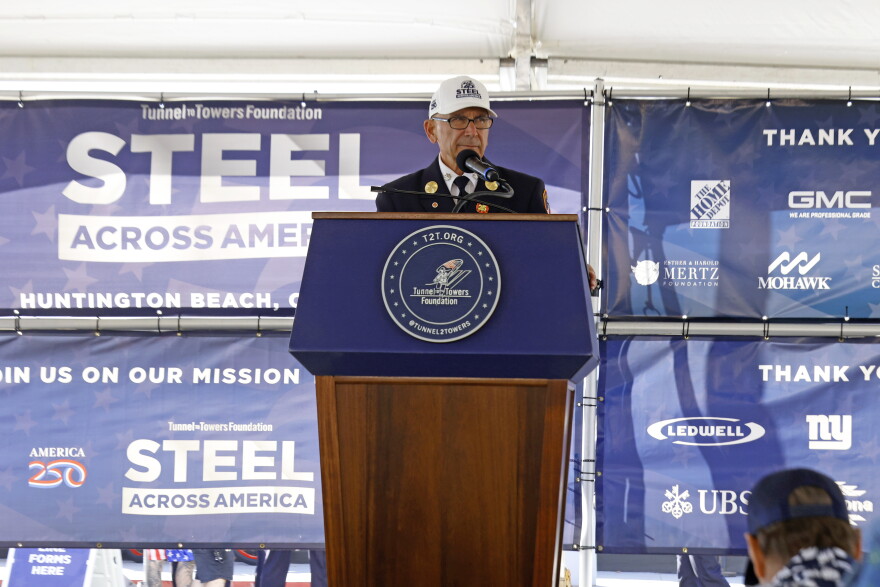 A man in a blue suit and white hat speaks at a podium in front of a sign that reads "Steel Across America."