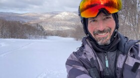 Man with beard and ski helmet standing on ski slopes with mountains in distance