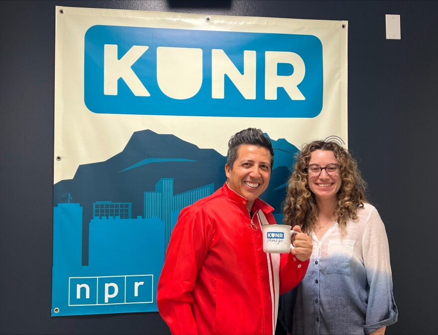 Two people are standing together in front of a banner with logos for KUNR and NPR. They are looking toward the camera while smiling. One is holding a KUNR Amigo mug.