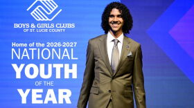 Young man in an olive suit holding a microphone on stage, beside text: “Boys & Girls Clubs of St. Lucie County, Home of the 2026-2027 National Youth of the Year.”