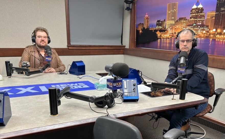 Two men wearing headphones sit at a table in a radio talk studio: the man at left has short brown hair and a brown mustache and beard, and is wearing glasses, a maroon patterned button-down shirt, and a brown jacket; the man at right has short dark hair and is wearing glasses, a dark blue sweater with a grey stripe, and jeans