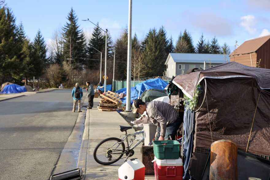 A few people standing along a sidewalk that has some small, tarp-roofed structures next to it.