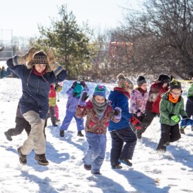 Large group of children bundled in winter clothing jumping and running in the show with an adult joining them