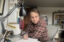 A man with short salt and pepper hair is wearing a plaid button-down shirt and drawing on a pad while sitting at a desk in an office.