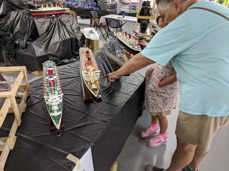 An older man points to a model ship with a young girl looking on.
