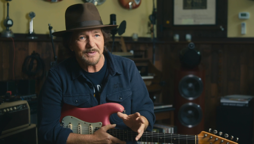 Eddie Vedder, wearing a brown felt fedora, sits in a studio holding a muted red stratocaster.