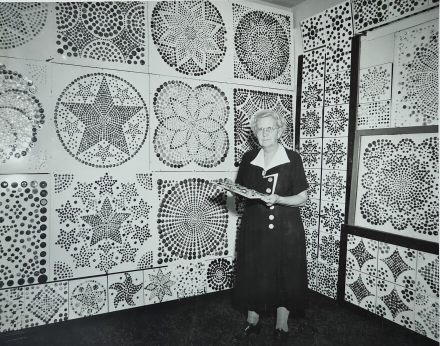 An old woman stands in a room filled with button designs and patterns on the walls