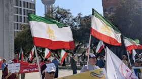 More than a dozen supporters of the National Council of Resistance of Iran (NCRI)  standing in JFK Plaza in Downtown Dallas, holding Iranian flags and signs. 