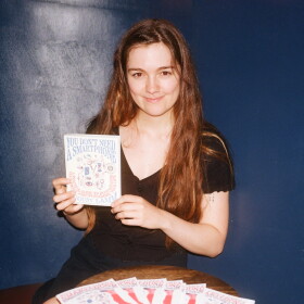 A woman with long brown hair holds a copy of the book "You Don't Need a Smartphone." Several copies of the book are splayed out on a table in front of her. 