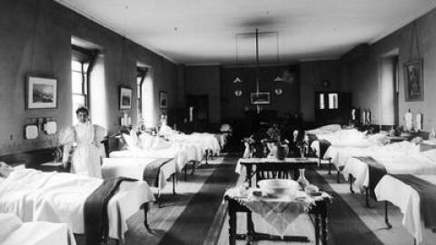 A black-and-white photograph shows a Victorian-era hospital ward. The room features two rows of neat iron beds with white linens and dark blankets at the foot. Patients are visible in several beds, while a nurse in a crisp white uniform with puffed sleeves stands near the window. In the center aisle, two wooden tables hold medical supplies, including a large white basin. 