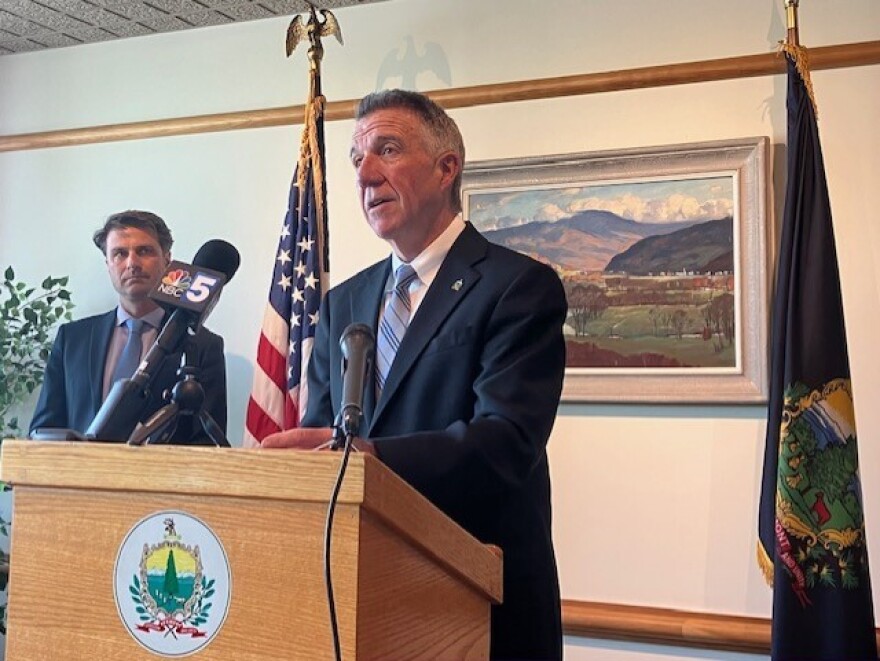 A man in a suit speaks at a wooden podium. Behind him is a landscape painting and an American flag and a Vermont flag.