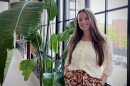Kristen Plonsky wears an off-white shirt and patterned brown pants while she smiles for a photo beside a large green plant and in front of a black-paned window. Plonsky worked as a Health Hub Coordinator with Detroit Public Schools Community District. After the federal government increased immigration enforcement, her role shifted to helping local families navigate the immigration system and supporting them with basic needs.