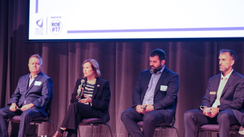 Four people sit in chairs on a stage. One is speaking into a microphone.