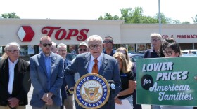 More than a dozen supporters stand alongside NY US Senator Chuck Schumer outside of a Tops grocery store in Auburn. One woman stands off to the side holding a green, white, and black sign reading, "Lower Prices for American Families."