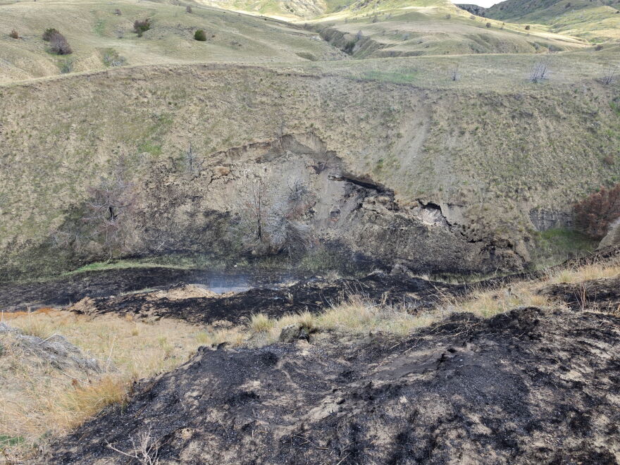 A wildfire caused by an exposed burning coal seam from May of 2025, image depicts blackened and smoking grass near the exposed seam.