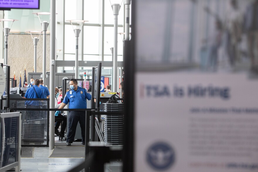 A TSA officer helps screen passengers at Austin-Bergstrom International Airport. A blurred out "TSA is hiring" sign appears in the foreground.