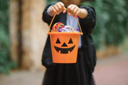 Little cute girl in witch costume holding jack-o-lantern pumpkin bucket with candies and sweets. Kid trick or treating in Halloween holiday