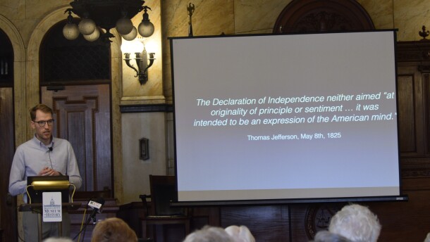 A tall Caucasian man stands at a podium. He's wearing a blue button-down shirt, and his hands are clasped in front of him. He's talking to a seated crowd. Next to the man is a screen that reads "The Declaration of Independence neither aimed "at originality of principle or sentiment ... it was intended to be an expression o f the American mind." -- Thomas Jefferson, May 8th, 1825