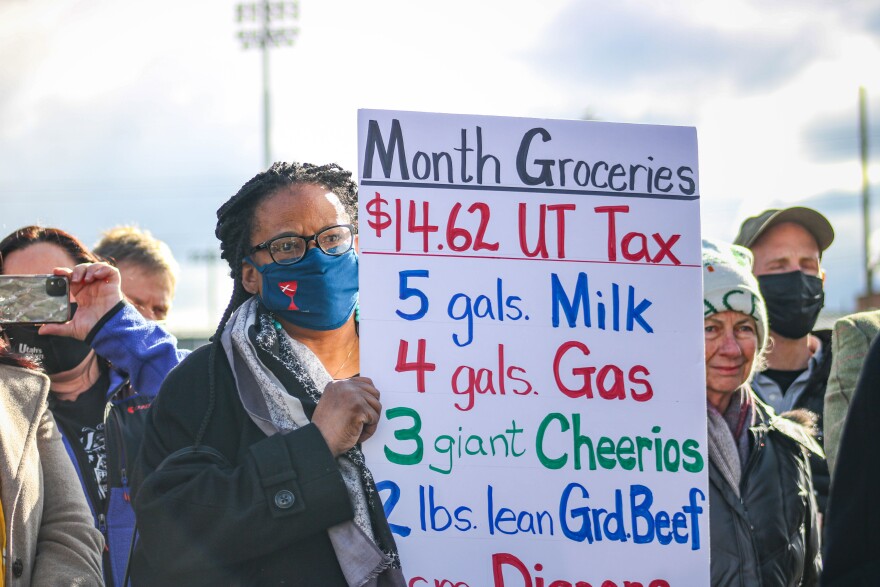 A photo of a Black woman in a face mask holding a sign that says 'Month Groceries' with a list of items and $14.62 total Utah Sales Tax. 