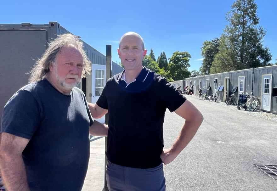 Two middle-aged men are standing next to each other outdoors. Behind them are shipping container homes. 
