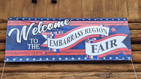 Red, white, and blue signage reading "Welcome to the Embarrass Region Fair" against a log wall