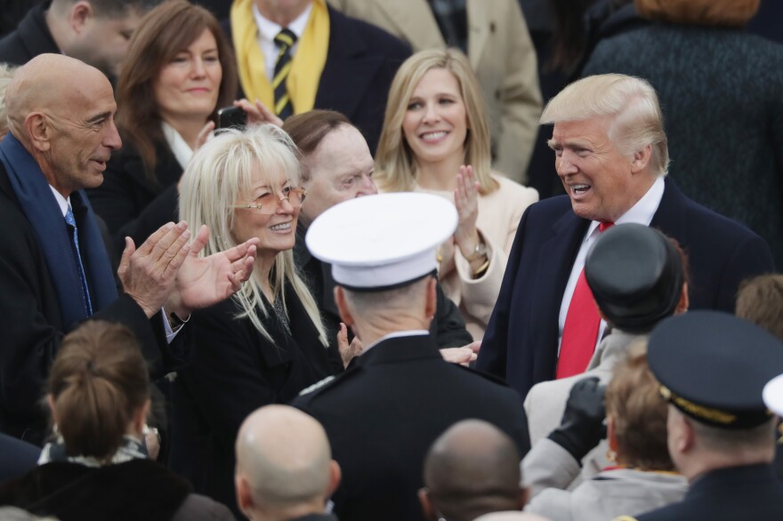 People stand close together in a group around former President Trump. They wear formal clothes and Tom Barrack claps.