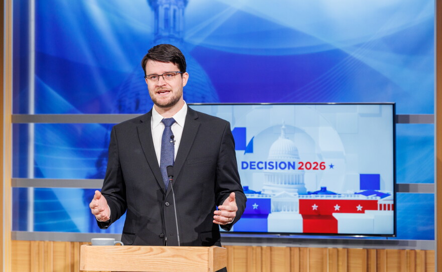 A man in a suit speaks at a podium in front of a TV that reads "Decision 2026"