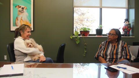 Two women sit at a large table in a board room. One holds a small, scruffy dog in her lap. Behind her is a framed photo of another dog. 