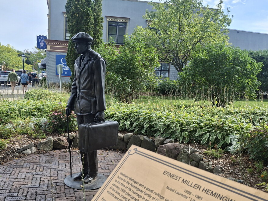 A statue of a young man with a suitcase and cane is visible in a park in downtown Petoskey. In the foreground, a gold plaque reads "Ernest Miller Hemingway." 