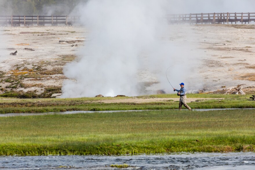 A man walks across a river landscape casting a fishing rod into the water as a geyser explodes behind him.